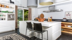 Una cocina moderna con una isla central, taburetes de bar, una cafetera y vistas al exterior con vegetación.