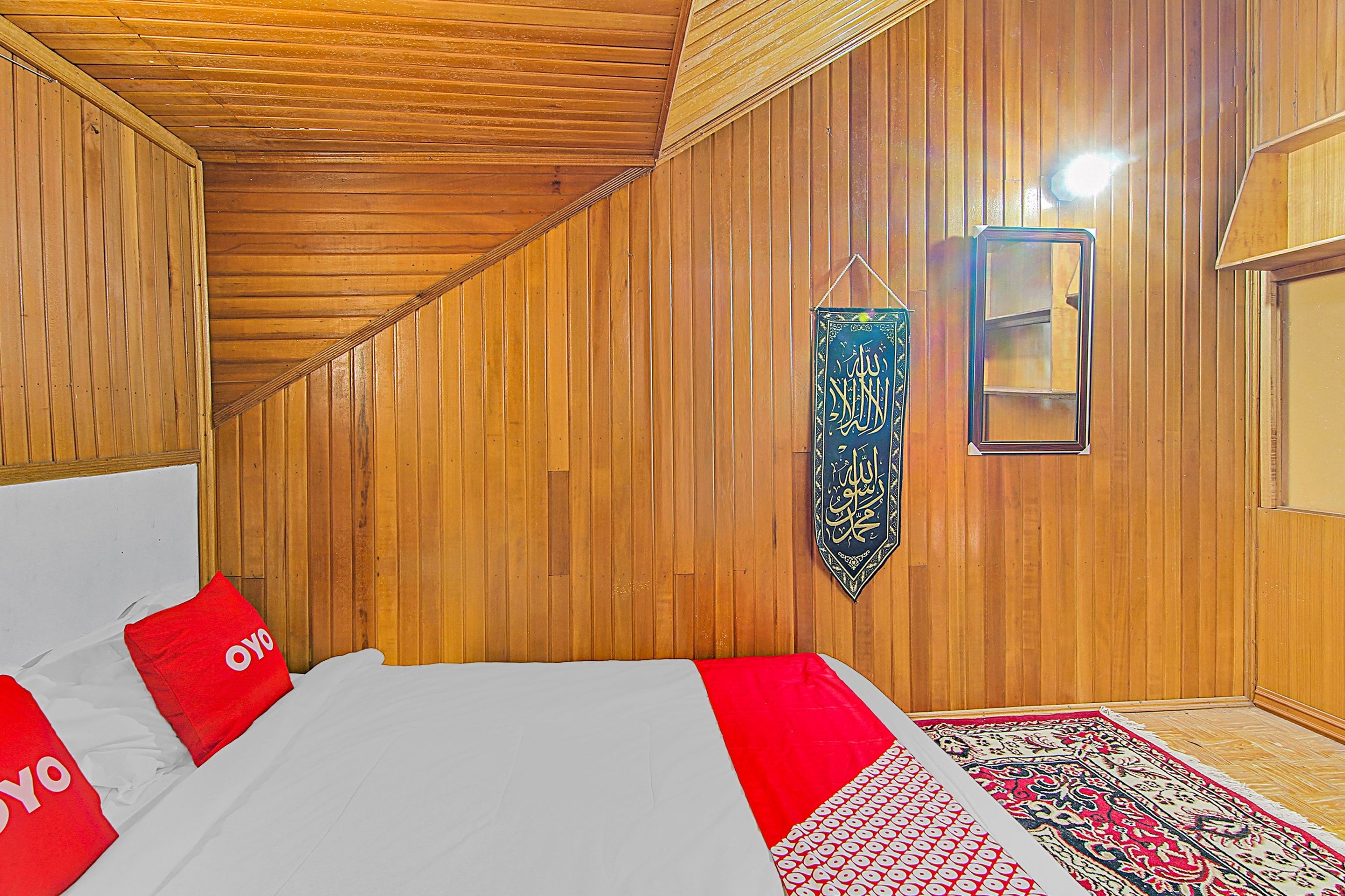 A room with wooden panel walls, a bed with red pillows, a hanging calligraphy piece, and a patterned rug.