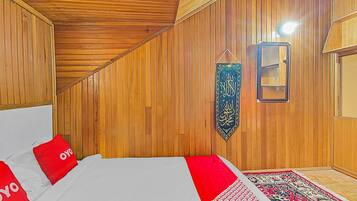 A room with wooden panel walls, a bed with red pillows, a hanging calligraphy piece, and a patterned rug.
