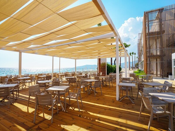 Un espacio de comedor al aire libre con mesas y sillas de madera, una terraza de madera y vista al mar.