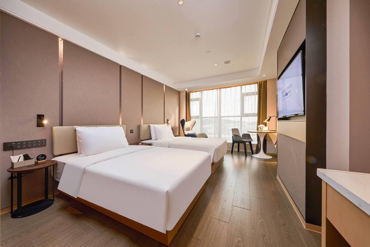 Une chambre d’hôtel avec deux lits, un bureau, une chaise, une télévision et une grande fenêtre.
