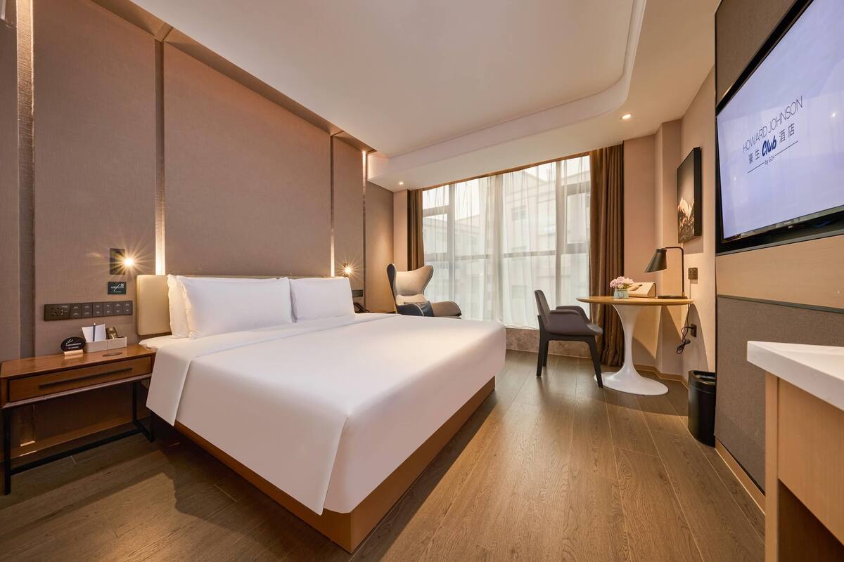 Une chambre d’hôtel avec un grand lit, un bureau, une chaise, une télévision et une fenêtre avec des rideaux.