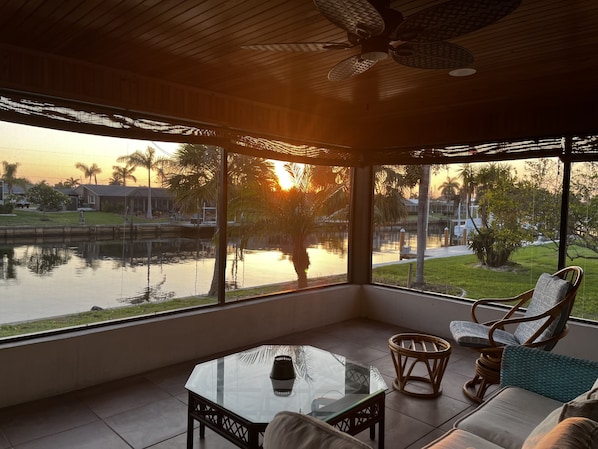 A covered outdoor seating area with a glass table, wicker chairs, and a view of a sunset over a body of water.