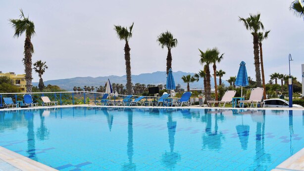 Ein Swimmingpool mit Liegestühlen und Sonnenschirmen, umgeben von Palmen und Bergen im Hintergrund.