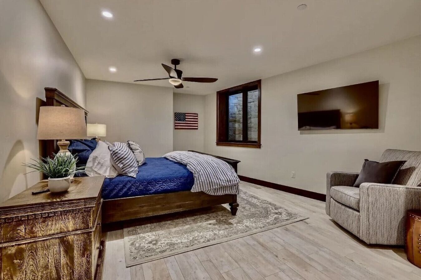 A bedroom with a bed, a desk, a chair, a ceiling fan, an American flag, and a TV on the wall.