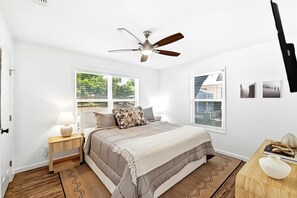 Quarto com cama, mesa de cabeceira, luminária, ventilador de teto, janelas e televisão fixada na parede.