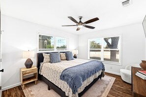 Un dormitorio con cama, mesitas de noche, ventilador de techo y ventanas con persianas.
