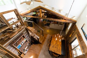 Een ruime loft-achtige kamer met een hoge plafond, houten balken, een eethoek met een lange tafel, en een keuken op de achtergrond.