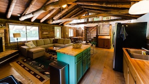 Interno di un rustico chalet con soffitto in legno, un accogliente soggiorno con divano e tavolo da pranzo in legno, una cucina con lavello e frigorifero, e un'ampia finestra che offre luce naturale.