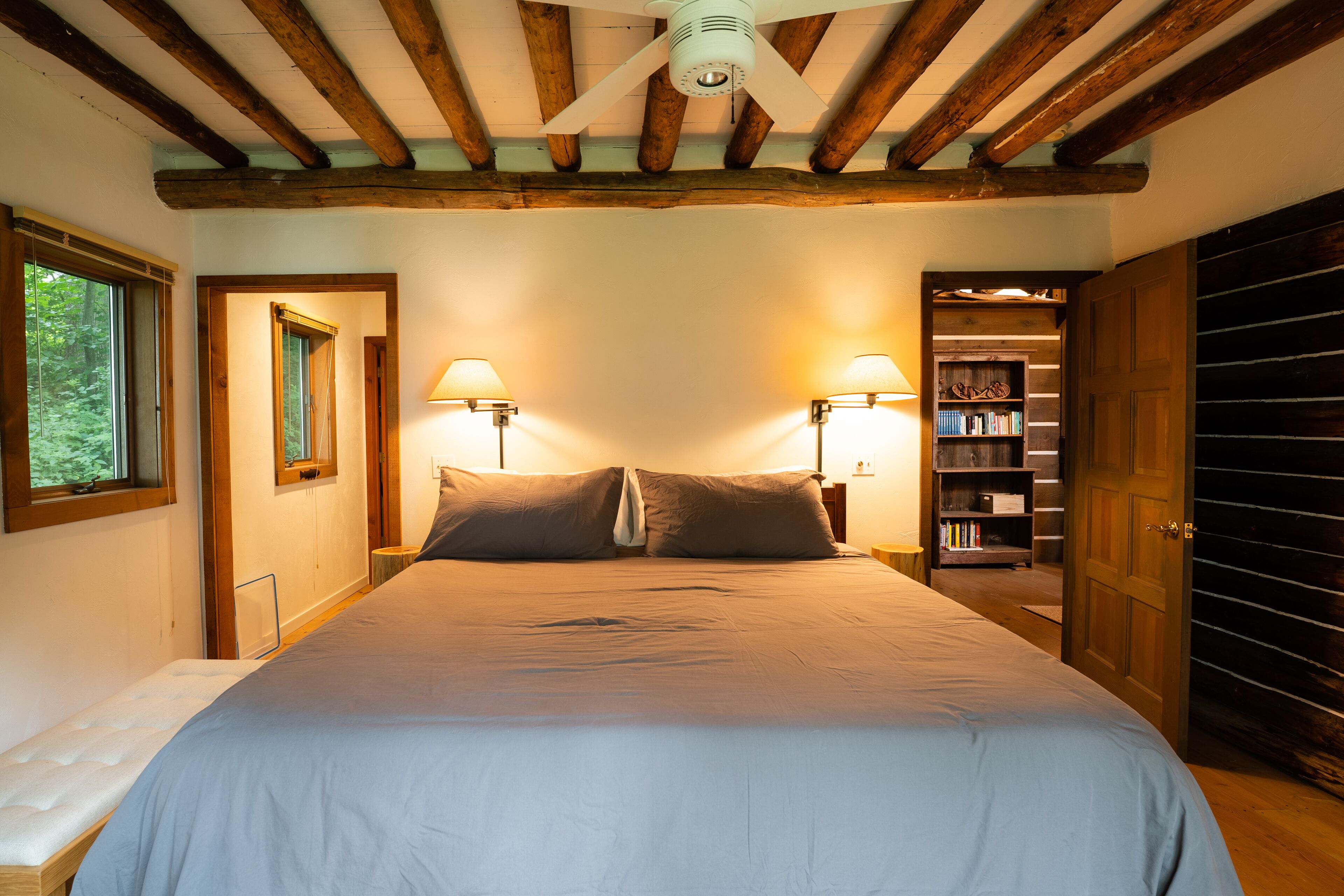 A bedroom with a large bed, wooden ceiling beams, two wall-mounted lamps, and a bookshelf.