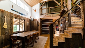 Un interno rustico con un tavolo e sedie di legno, una scala con ringhiera in legno e una testa di cervo appesa al muro.