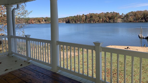 A covered porch with white railings overlooking a lake, with trees and a distant house visible.