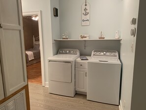 Ein Waschraum mit zwei weißen, frontbeladenen Waschmaschinen und Trocknern, einem kleinen Regal mit Dekoration und einer Tür zu einem Schlafzimmer.