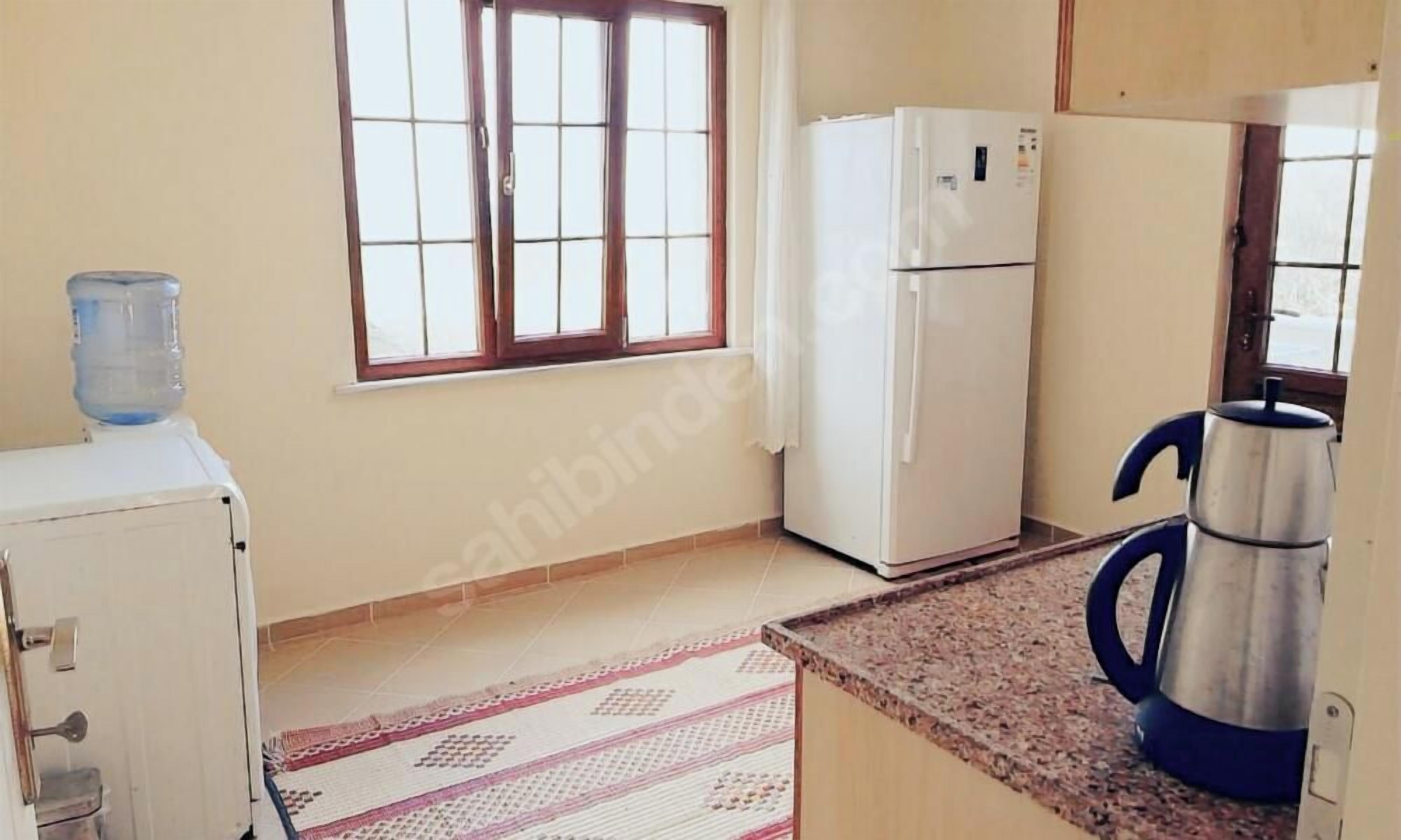 A kitchen with a refrigerator, a water dispenser, a kettle, and a window with curtains.