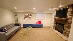 A living room with a grey sofa, a blue sofa with red cushions, a glass coffee table, a stone fireplace, and a mounted TV. - Entire Modern Basement Apt in Sandy—1 min to I-15 (Sandy)