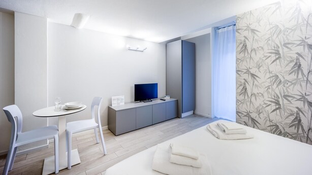 A modern hotel room with a bed, a desk with a TV, a chair, and a small table with glasses and a bowl.