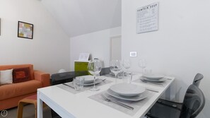 Ein Essbereich mit einem weißen Tisch, gedeckt für vier Personen, Gläsern, Tellern und Besteck. Im Hintergrund sind ein grüner Schrank und eine weiße Tür zu sehen.