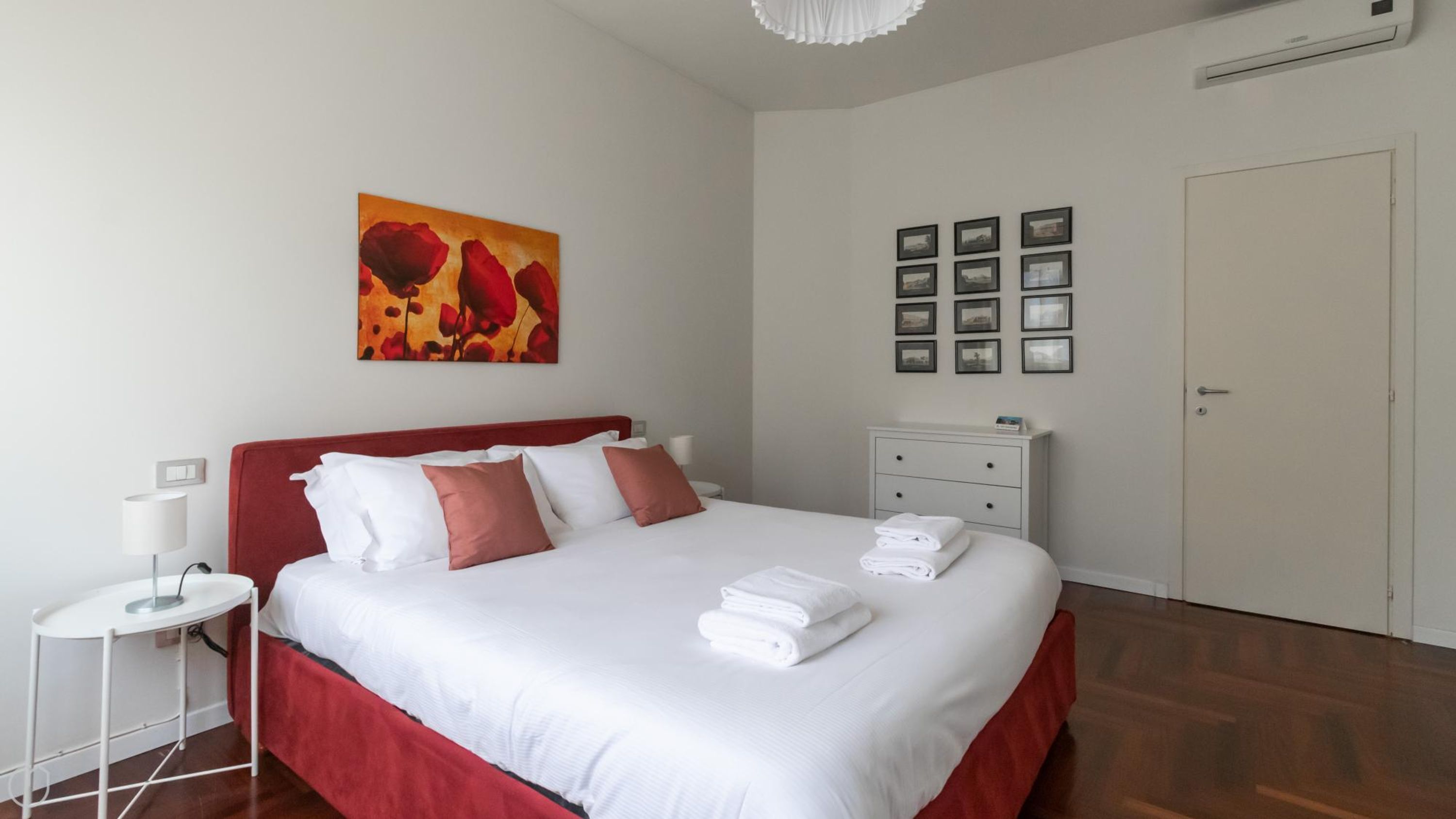 Ein ordentlich bezogenes Bett mit weißer und roter Bettwäsche, ein roter Kopfteil und ein kleiner Nachttisch mit einer Lampe.