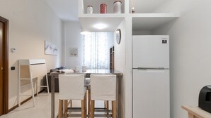 A compact kitchen with a refrigerator, a dining table with chairs, and a small shelf with decorative jars. - Italianway Easy - Pisino 5 (Milan)