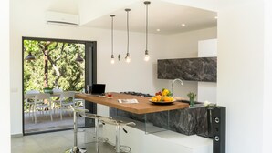 A modern kitchen with a central island, bar stools, and a dining area with a view of trees. - Villa Charme 6 in Pollina (Pollina)