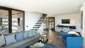 Uma sala de estar moderna com um sofá cinzento, uma poltrona azul e uma mesa de centro de vidro. Uma escadaria dá acesso a um andar superior.