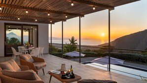 Um terraço com móveis de vime, uma mesa de jantar com copos de vinho e vista para o pôr do sol sobre o mar.