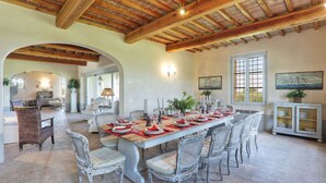 A dining area with a long table set for a meal, surrounded by wicker chairs, under a wooden ceiling with exposed beams.