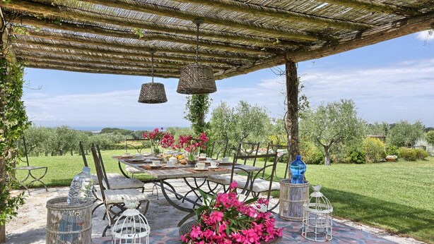 An outdoor dining area with a table set for two, surrounded by greenery and a view of the landscape.