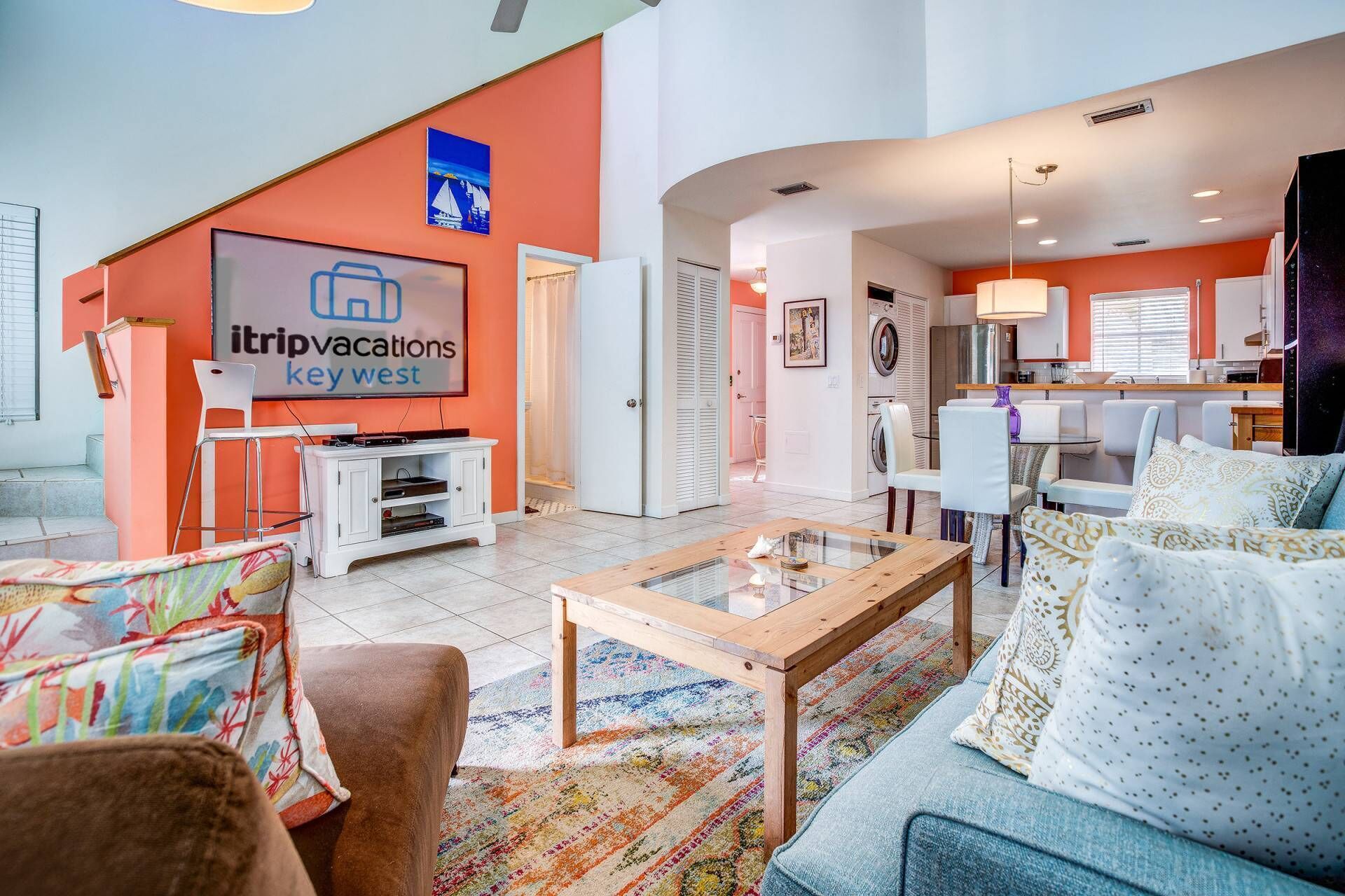 A modern living room with a flat-screen TV, a coffee table, and a sofa. There is a kitchen area in the background with a dining table and chairs.
