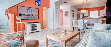 A modern living room with a flat-screen TV, a coffee table, and a sofa. There is a kitchen area in the background with a dining table and chairs.