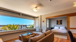 A modern hotel room with a large balcony, a bed, a sofa, and a desk with a lamp. - The Sands - Apartment 12 Onetangi Beach (Waiheke Island)