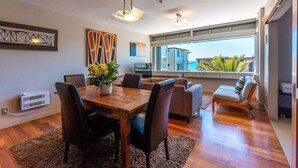 A modern dining area with a wooden table and chairs, a potted plant, and a view of the sea through large windows. - The Sands - Apartment 12 Onetangi Beach (Waiheke Island)