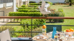 Un balcón con una mesa lista para el desayuno, que incluye bollería, frutas y bebidas. Una gaviota está posada en la barandilla.