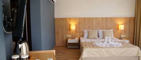 Habitación de hotel con cabecera de madera, dos lámparas de noche, cama decorada con un cisne y pétalos de rosa, mesita de noche de madera con un teléfono y una cafetera sobre una bandeja.