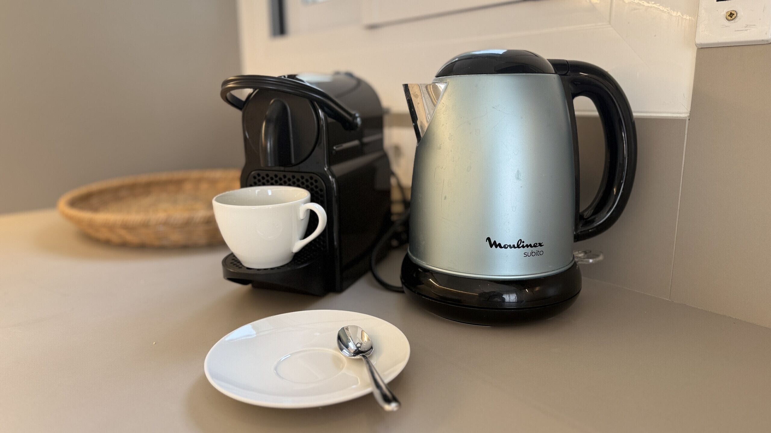 A coffee maker, a cup, and a plate with a spoon on a kitchen counter.