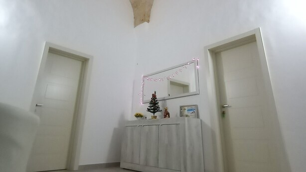 Una habitación con techo abovedado alto, paredes blancas y piso de baldosas. Se observan dos puertas, una mesa pequeña con un espejo y algunos objetos decorativos.