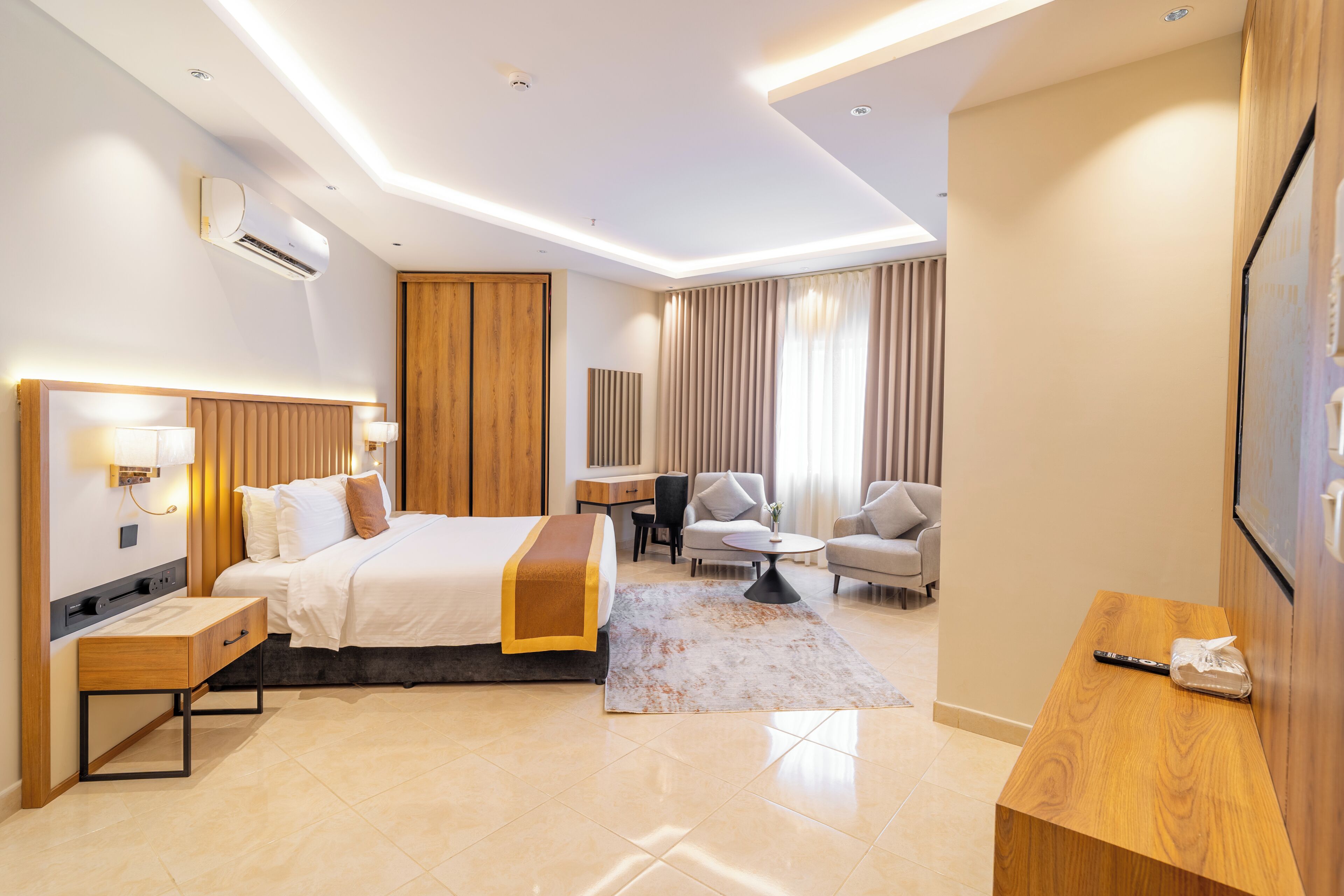 A modern hotel room with a bed, bedside tables, a desk, a chair, a sofa, and a TV.