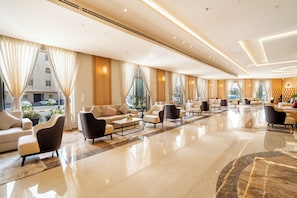A spacious hotel lobby with a polished marble floor, multiple seating areas, and large windows offering a view of the street.