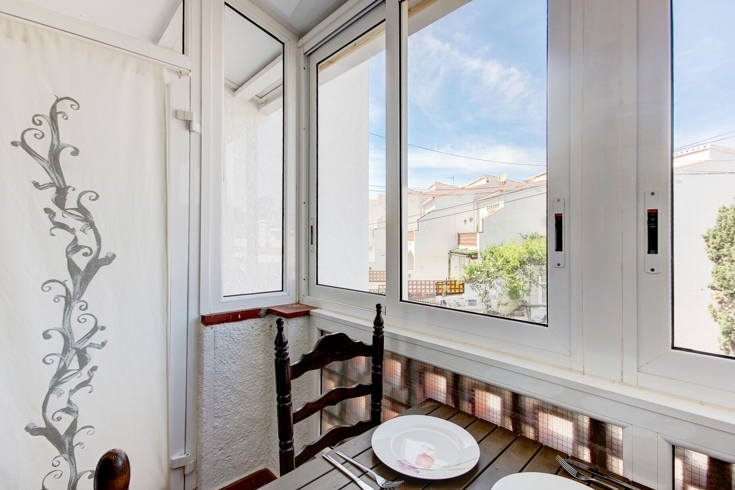 A room with a large window, a wooden chair, a dining table with a plate and cutlery, and a decorative wall design.