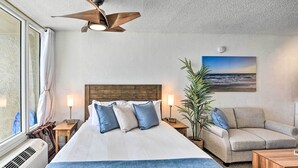 A hotel room with a bed, a sofa, a ceiling fan, a nightstand with a lamp, a potted plant, and a painting of a beach scene. - Oceanfront, Fourth-Floor Condo on Daytona Beach! (Daytona Beach)