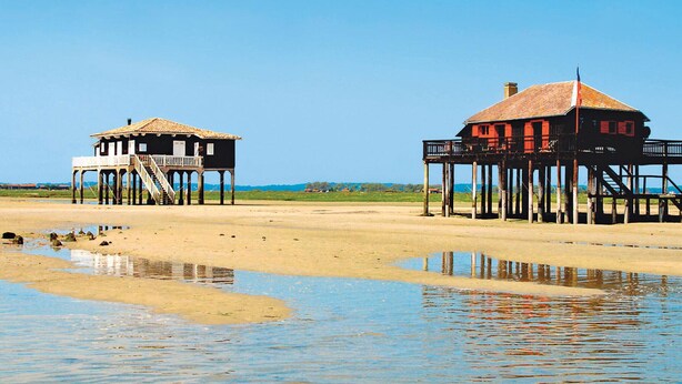 Two wooden structures with balconies, one with a staircase and the other with a flat roof, on a sandy beach with calm water in the foreground. - Central Arcachon: Modern 2BR Apartment with Terrace, 400m from Beach (Arcachon)