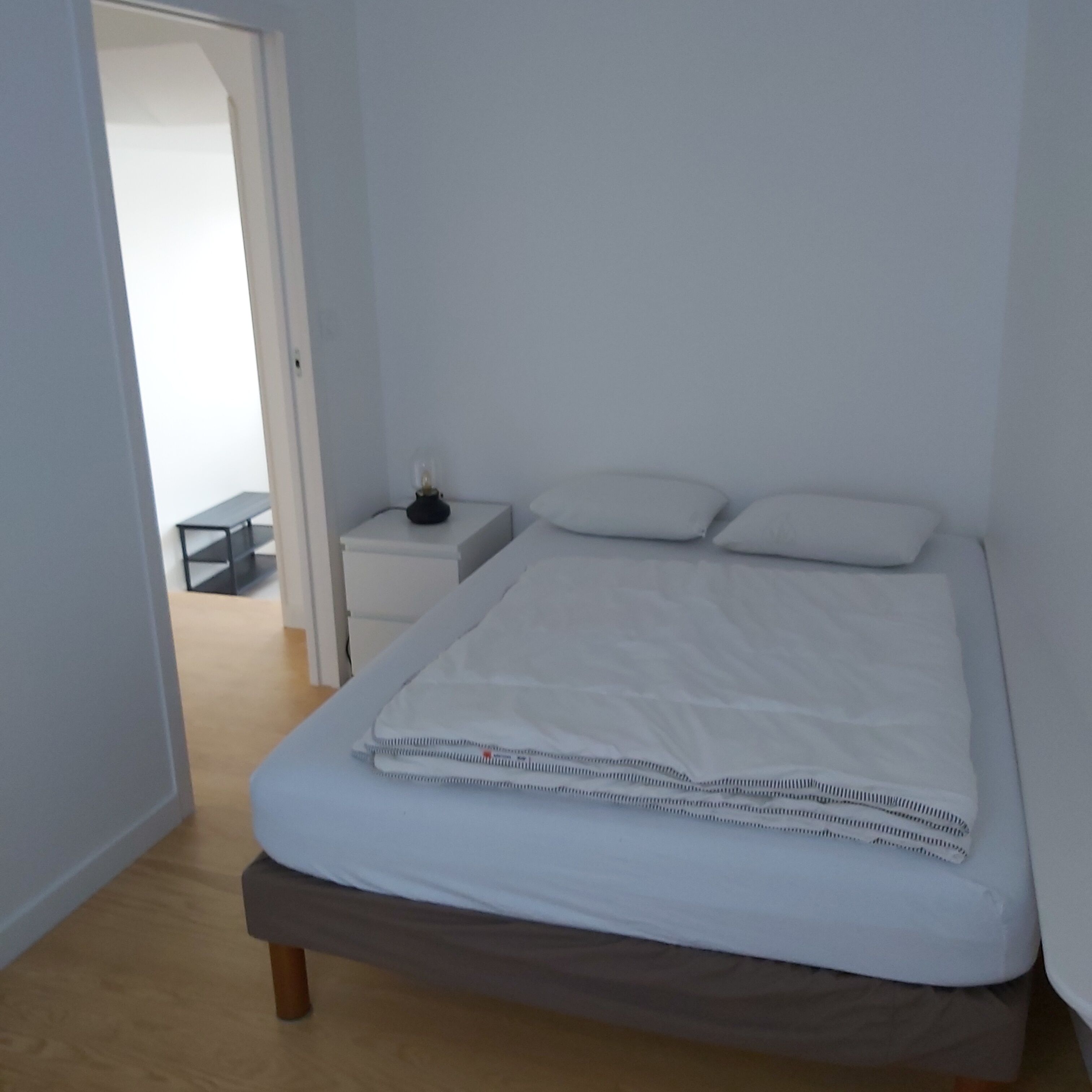 Une chambre avec un lit simple, une table de chevet avec une lampe et un petit banc dans un coin.