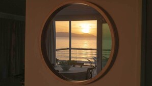 Ein rundes Fenster mit Blick auf einen Sonnenuntergang über dem Wasser, ein Tisch und eine Bank.