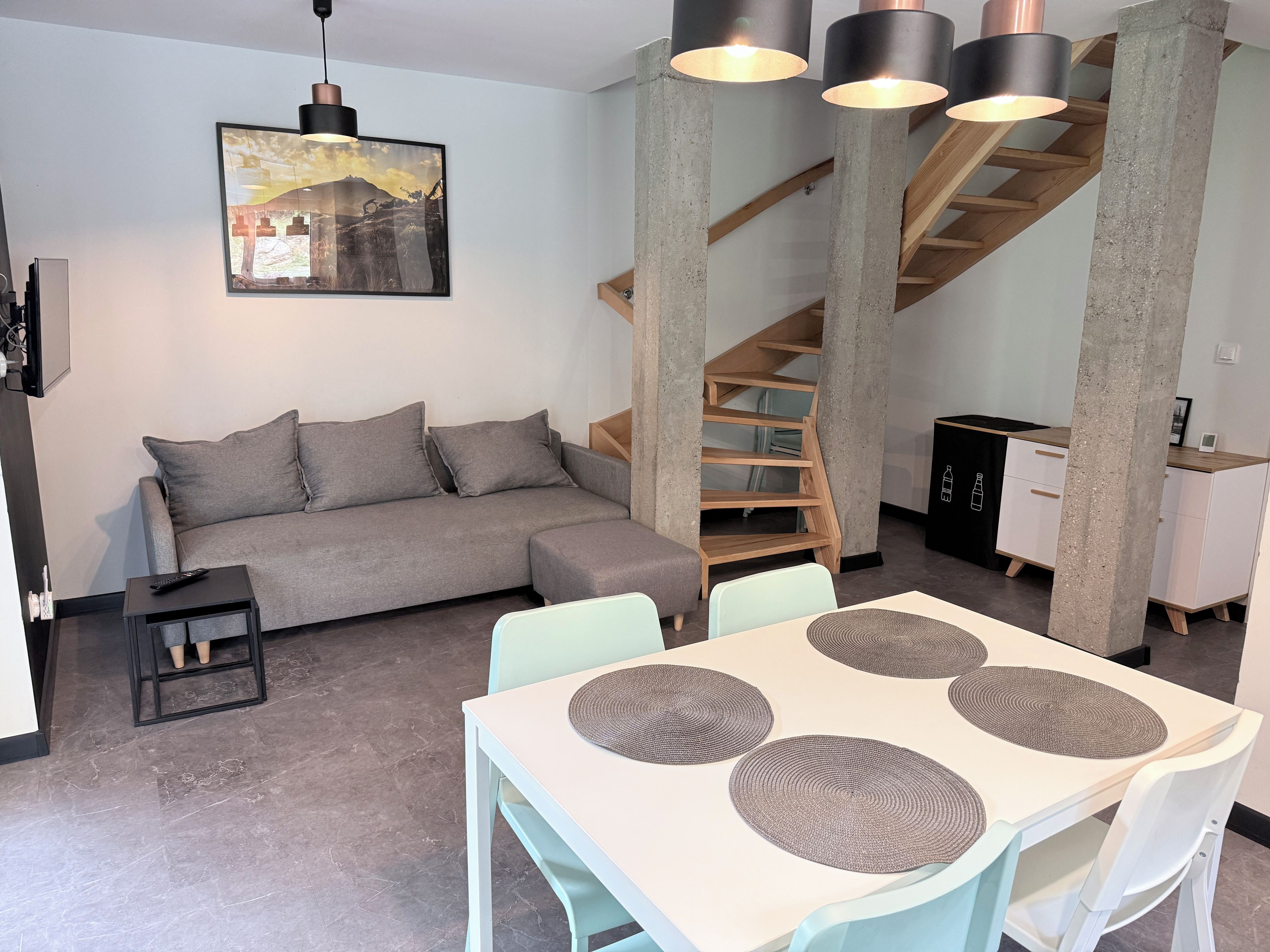 A modern living space with a grey sofa, a dining table with placemats, a staircase, and a hanging light fixture.