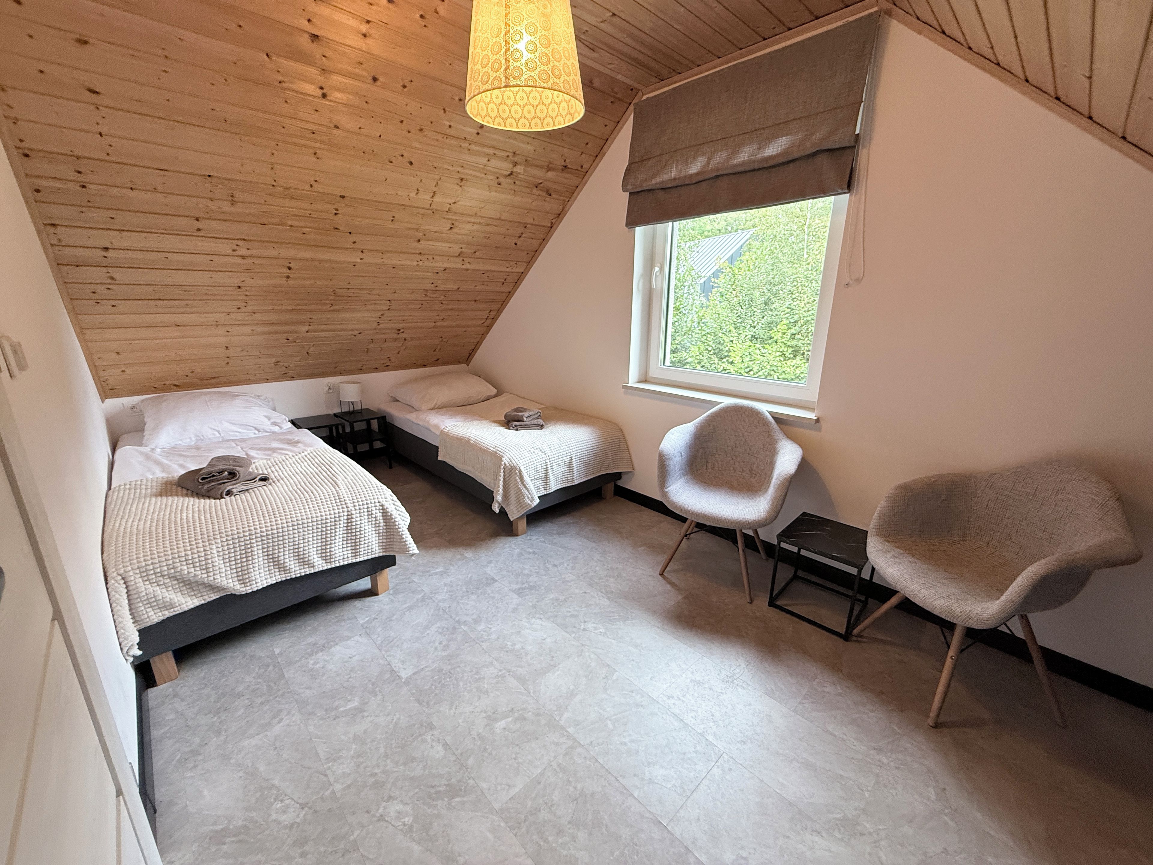 A bedroom with two beds, a window with a view, a chair, and a hanging light fixture.
