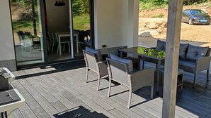 Un patio exterior con mobiliario de mimbre, una puerta de cristal que da a otra zona de patio, y una vista de un coche y el paisaje al fondo.