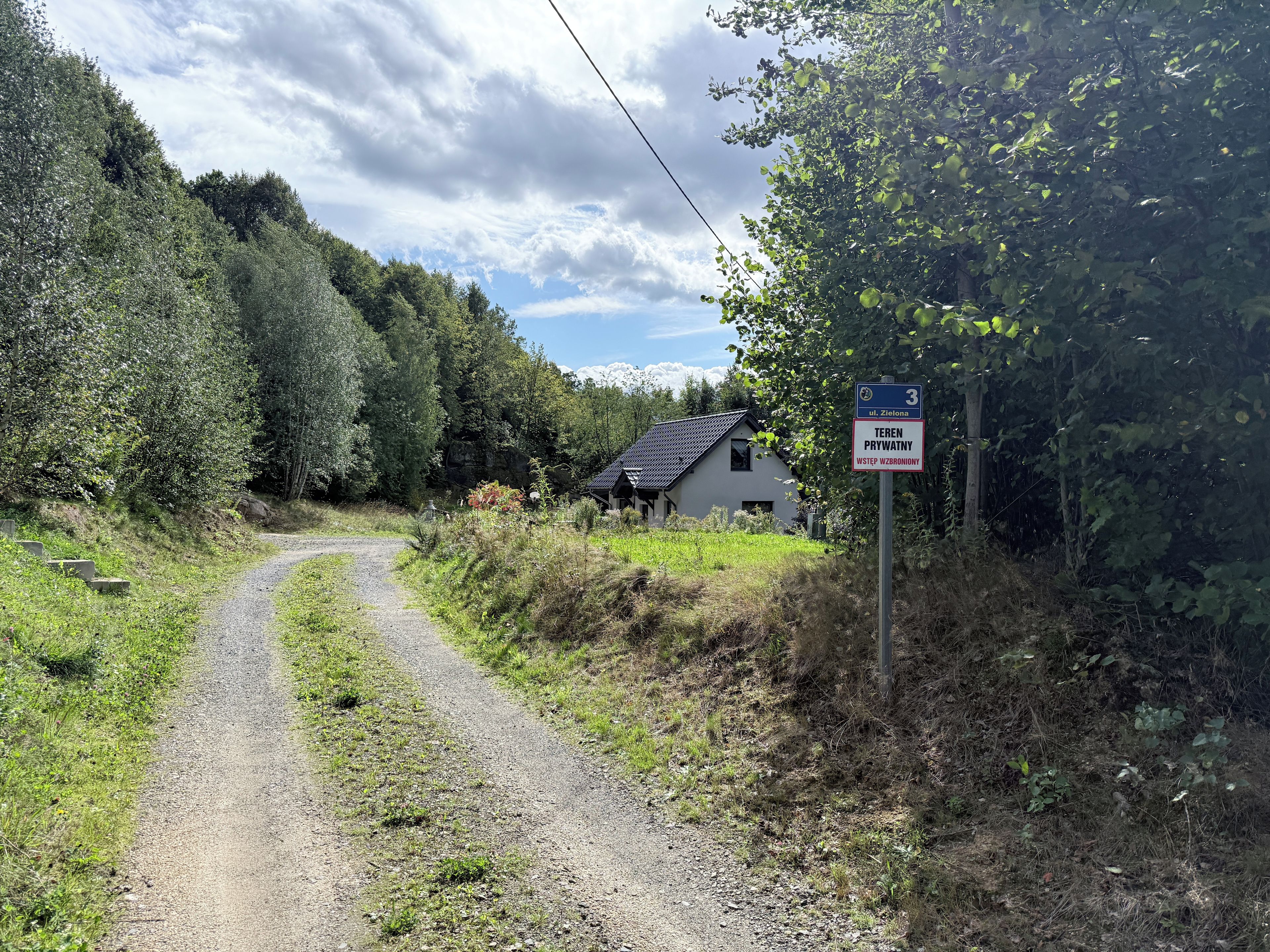 Un chemin de campagne mène à une maison blanche au toit sombre, entourée d’une végétation luxuriante et sous un ciel nuageux.