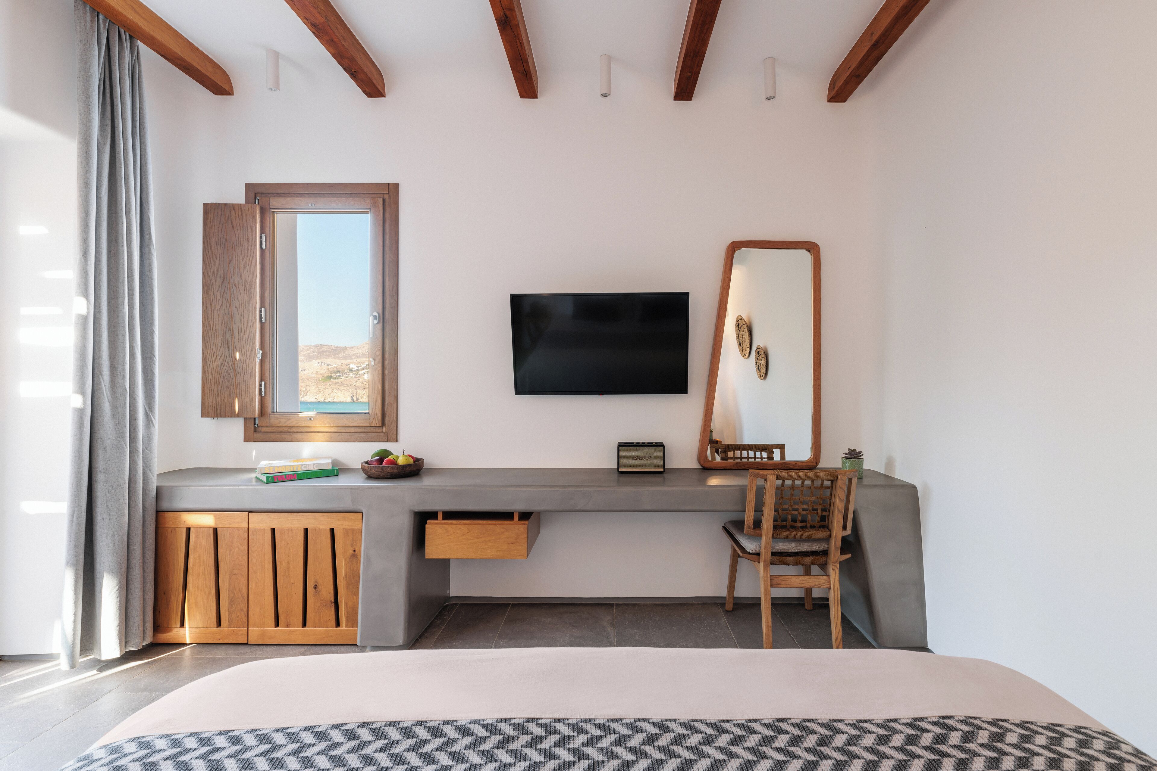 A bedroom with a bed, a desk with a TV, a mirror, and a window with a view.