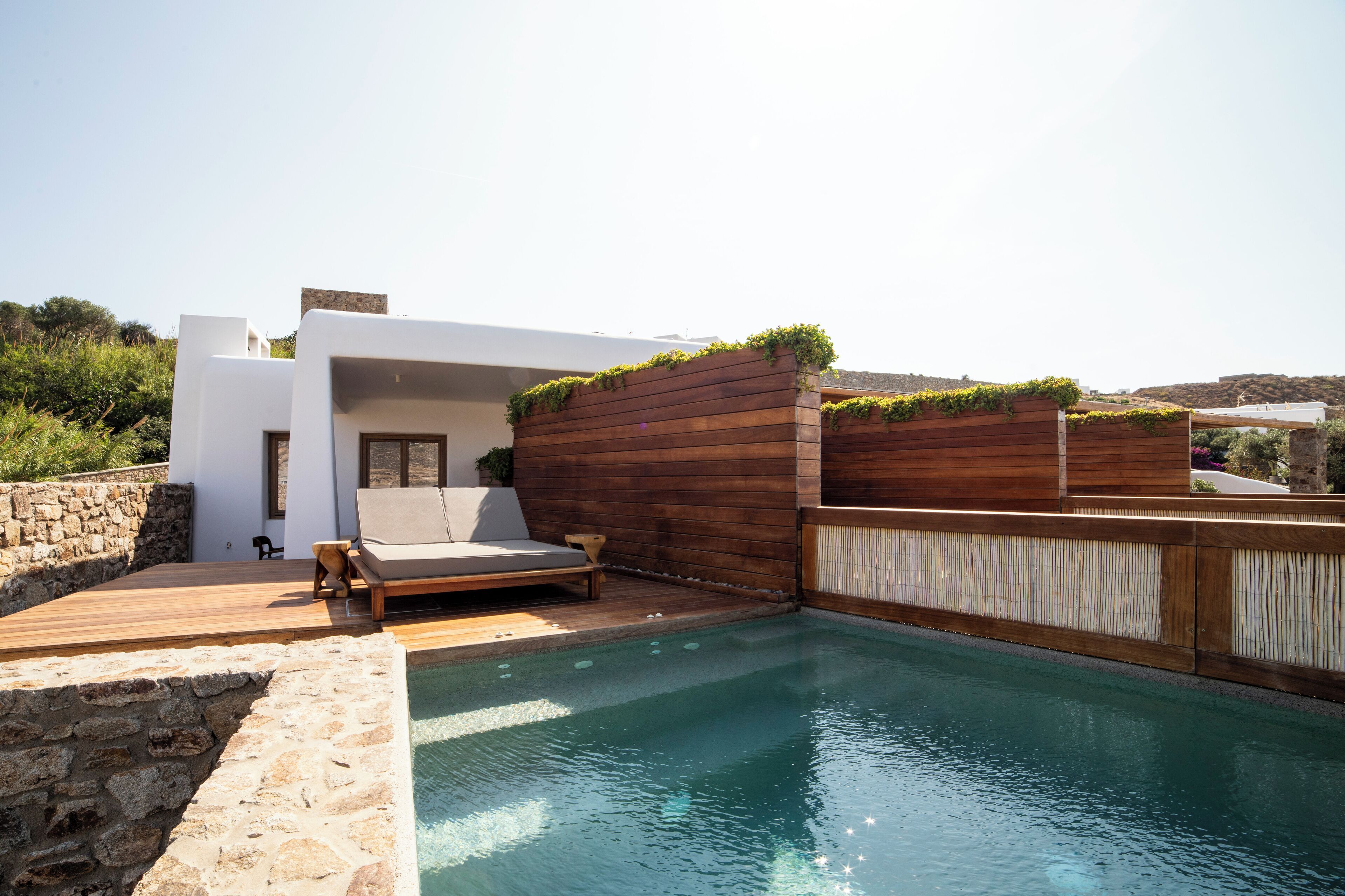 Area piscina moderna all'aperto con un solarium in legno, una poltrona da relax e un muro in pietra.
