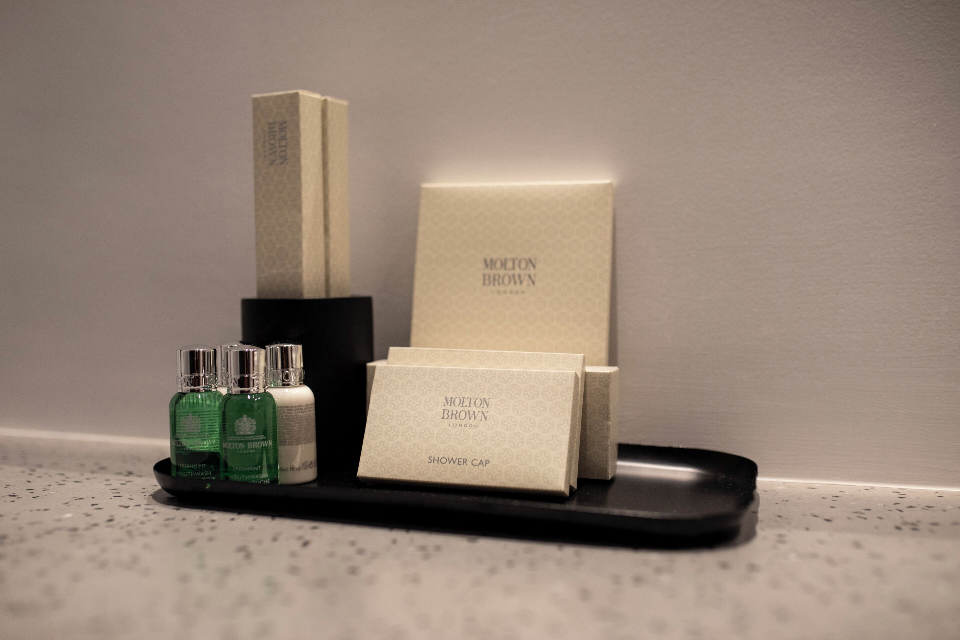 A tray with a shower cap, a box, and two bottles of liquid on a speckled countertop.